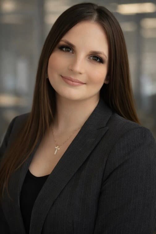 Smiling woman with long brown hair wearing a pinstripe blazer and a small cross necklace.